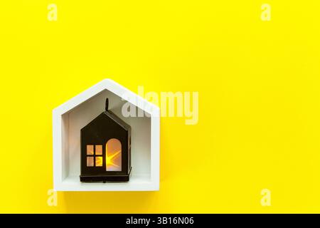 Piccola lanterna nera a forma di casa con una luce calda all'interno posta in una cornice bianca a forma di casa, posta su uno sfondo giallo brillante in un accogliente e moderno Foto Stock