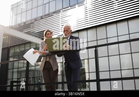 Due colleghi che si appoggiano sulla recinzione all'esterno dell'edificio ofiice che parlano e guardano un notebook Foto Stock