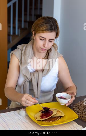 Donna che distribuisce marmellata di frutti di bosco su pancake a colazione. Giovane donna con un vestito casual che diffonde marmellata di frutti di bosco su pancake con un cucchiaio, gustando un piatto fatto in casa Foto Stock