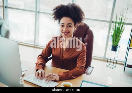 Giovane donna d'affari con capelli naturali alla sua scrivania in un ufficio moderno, sorridente e lavora al computer in piena luce naturale Foto Stock