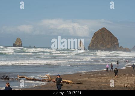 CANNON BEACH, OREGON, USA - 12 aprile 2025: I visitatori camminano lungo le spiagge sabbiose di Cannon Beach sotto un cielo nuvoloso, con l'iconica Haystack Rock in piedi. Foto Stock
