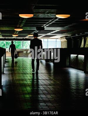 Due uomini camminano attraverso un sottopassaggio moderno e poco illuminato, le loro silhouette contrastano con il fogliame verde brillante visibile attraverso una finestra. Foto Stock