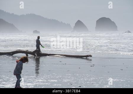 CANNON BEACH, OREGON, USA - 12 aprile 2025: I visitatori camminano lungo le spiagge sabbiose di Cannon Beach sotto un cielo nuvoloso, con l'iconica Haystack Rock in piedi. Foto Stock
