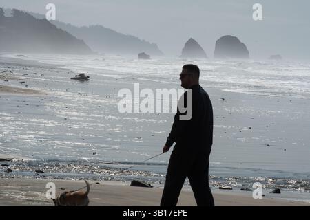 CANNON BEACH, OREGON, USA - 12 aprile 2025: I visitatori camminano lungo le spiagge sabbiose di Cannon Beach sotto un cielo nuvoloso, con l'iconica Haystack Rock in piedi. Foto Stock