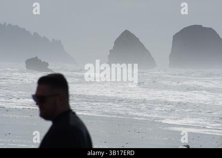CANNON BEACH, OREGON, USA - 12 aprile 2025: I visitatori camminano lungo le spiagge sabbiose di Cannon Beach sotto un cielo nuvoloso, con l'iconica Haystack Rock in piedi. Foto Stock