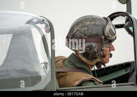 Pilota con casco e occhiali di protezione in pelle vintage siede nella cabina di pilotaggio di uno Spitfire RAF, goccioline sul baldacchino che accennano alla recente pioggia a Duxford, in Inghilterra. Foto Stock