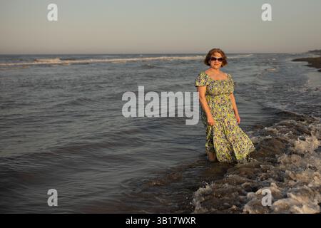 Donna anziana in piedi al mare la sera. Indossa un maxi vestito floreale. L'atmosfera generale dell'immagine è tranquilla e serena, Foto Stock