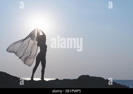 Una ballerina ballerina con un mantello trasparente in mano, che fluttua nel vento. Silhouette di una donna danzante in riva al mare contro il cielo, un empio Foto Stock