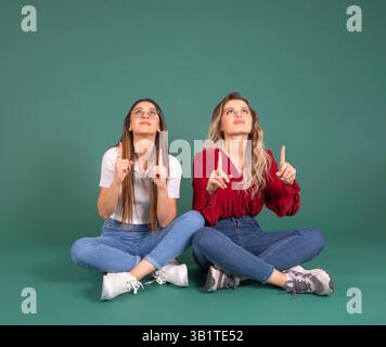 Looking and pointing up, full size two charming adorable caucasian women sitting crossed legs looking and pointing up mock up advertisement concept Foto Stock