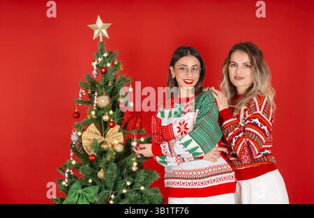 Due belle donne caucasiche che posano vicino all'albero di Natale decorato. Vigilia di Capodanno. Divertirsi, preparare le feste, festeggiare indossare maglioni Foto Stock