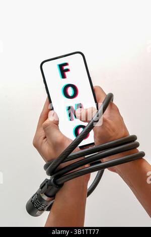 Le mani sono bloccate con un blocco cavo, tenendo in mano uno smartphone con FOMO (paura di perdere): Dipendenza dai social media, paura di perdere notizie Foto Stock