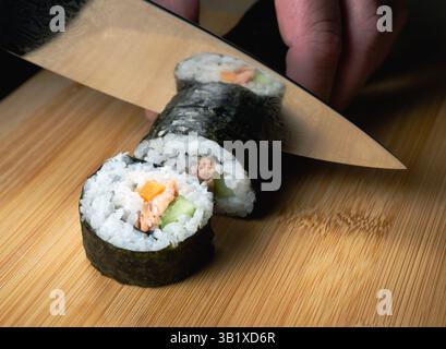 Una persona sta tagliando un sushi roll con un coltello. Il sushi roll è composto da riso, salmone e verdure. Il coltello è posizionato al centro dello sterzo Foto Stock