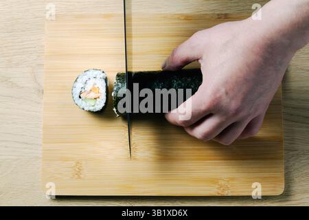 Una persona sta tagliando un rotolo di sushi su un tagliere di legno. Il sushi roll ha un cetriolo di pesce e una carota all'interno. Foto Stock
