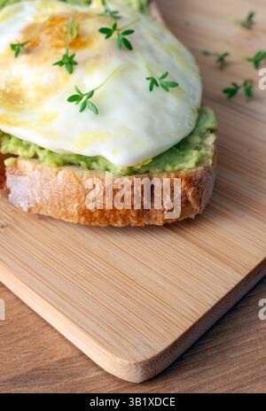 una fetta di pane tostato ricoperto da purè di avocado, un uovo fritto e fresche sorgenti verdi su un asse di legno Foto Stock