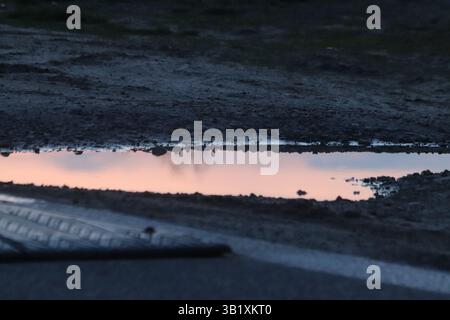 Una piccola pozzanghera circondata da sabbia e rocce con un riflesso del cielo al tramonto accanto a una strada con un dissuasore di velocità con erba sullo sfondo Foto Stock