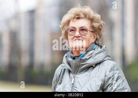 Ritratti una bella donna anziana con capelli corti e occhiali, in piedi all'aperto all'inizio della primavera, indossando una giacca calda, con un aspetto tranquillo, sicuro di sé e grazia Foto Stock