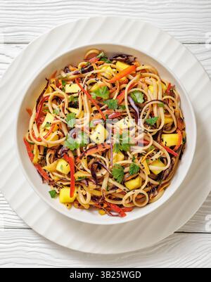 Insalata tailandese di tagliatelle di mango con pepe, cavolo rosso, menta e coriandolo, salsa di arachidi in una ciotola bianca su un tavolo di legno bianco, vista dall'alto verticale Foto Stock