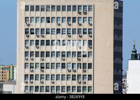 Vista ravvicinata della facciata di un vecchio edificio di uffici con finestre ripetitive e numerose unità di aria condizionata, sotto un cielo azzurro. Foto Stock