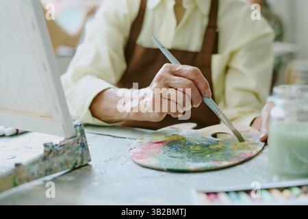 Primo piano medio di una donna anziana irriconoscibile che tiene il pennello mentre mescola le vernici a olio su una tavolozza di legno sporco Foto Stock