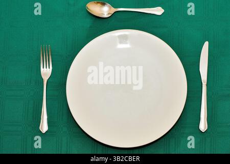 Vista ravvicinata di un tavolo con piatto bianco grande, coltello, forchetta e cucchiaio su una tovaglia verde scuro. Foto Stock