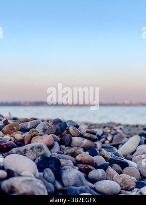 Primo piano di ciottoli da spiaggia con consistenza liscia e bassa profondità di campo, mare sfocato e cielo pastello sullo sfondo Foto Stock
