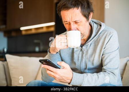 Uomo che beve caffè e usa il telefono cellulare a casa la mattina, attenzione selettiva Foto Stock