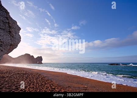 Il sole della mattina presto splende attraverso la foschia degli spruzzi, guardando indietro Durdle Door dalla spiaggia Foto Stock