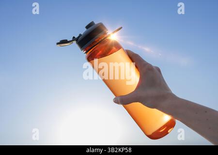 Primo piano della mano di una donna che tiene una bottiglia d'acqua riutilizzabile contro il cielo blu, concetto di idratazione, sostenibilità e stile di vita sano all'aperto. Foto Stock
