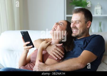 Coppia divertente che rideva insieme controllando il contenuto del telefono seduto su un divano a casa Foto Stock