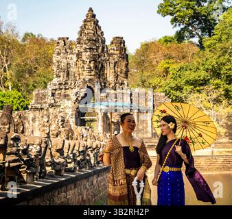 Due donne in abbigliamento tradizionale, con ombrelloni, stanno di fronte a un antico tempio con sculture in pietra e vegetazione lussureggiante. Foto Stock