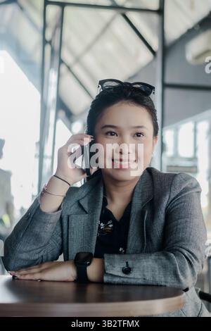 Donna d'affari asiatica sicura, con un abbigliamento elegante, gode di una conversazione telefonica allegra durante una pausa al caffè, mescolando svago e comunicazione moderna Foto Stock