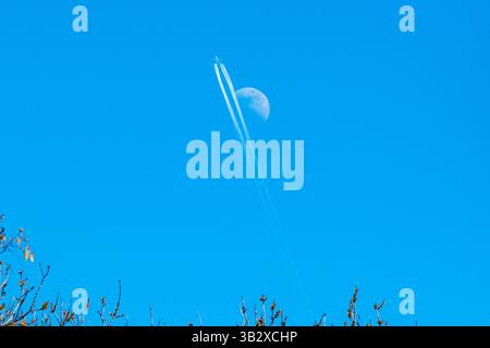 Un aereo in contrail passa il primo quarto di luna (4/5/25) in un cielo blu pomeridiano, Londra, Regno Unito Foto Stock