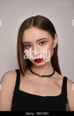 una giovane donna mostra un'espressione sicura indossando un rossetto rosso e un semplice top nero. I suoi lunghi capelli sono lisci, per esaltare la sua pelle Foto Stock