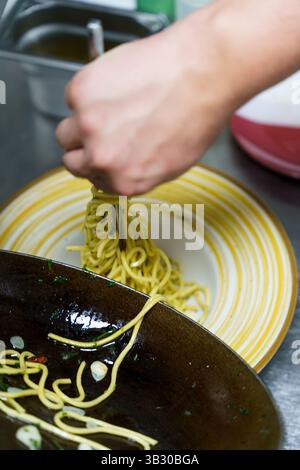 Uno chef prepara con cura una porzione di spaghetti aglio e olio utilizzando pinze in una cucina professionale. La pasta è ricoperta di olio d'oliva, aglio, prezzemolo, Foto Stock