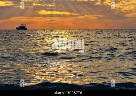 Riflesso del tramonto sulle dolci onde del mare con una barca lontana all'orizzonte. Tranquilla vista serale catturata dal livello dell'acqua. Foto Stock