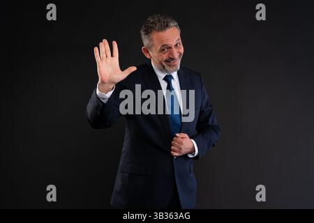 Allegro uomo d'affari in abito scuro e cravatta blu saluta con una mano alzata, sorridente in modo amichevole e accogliente, isolato su sfondo nero Foto Stock