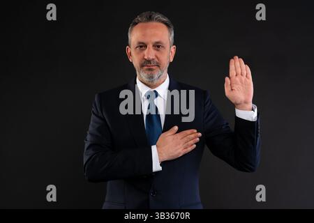 Uomo d'affari maturo in tuta che solleva la mano destra e mette la mano sinistra sul petto, simboleggia l'onestà, il giuramento o la promessa, su uno sfondo nero da studio. Foto Stock