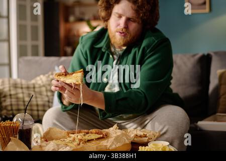 Uomo seduto su un divano nell'accogliente soggiorno che regala una fetta di pizza al formaggio con formaggio fuso che si estende dalla pizza. Bevande e spuntini sul tavolo accanto a lui per creare un'atmosfera informale Foto Stock