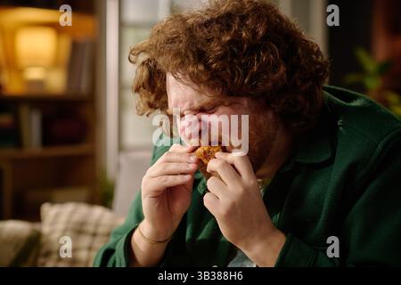 L'uomo assaggia pollo fritto croccante in una stanza accogliente e ben illuminata con scaffali e oggetti decorativi sullo sfondo Foto Stock