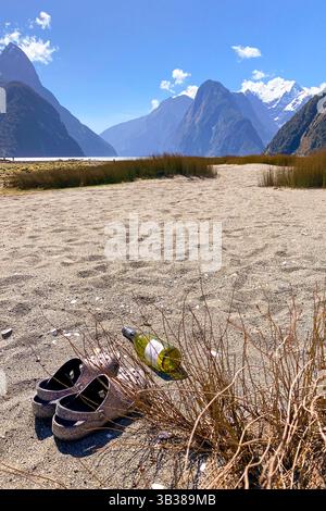 Scarpe abbandonate e una bottiglia di vino posta sulla sabbia nella baia di Milford Sound, un'area di bellezza naturale nei fiordi dell'Isola del Sud della nuova Zelanda Foto Stock