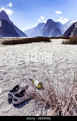 Scarpe abbandonate e una bottiglia di vino posta sulla sabbia nella baia di Milford Sound, un'area di bellezza naturale nei fiordi dell'Isola del Sud della nuova Zelanda Foto Stock