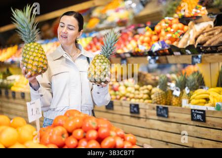 Donna positiva che sceglie gli ananas maturi nella sezione dei prodotti del supermercato Foto Stock