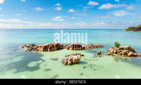 Enormi rocce granitiche riposano in acque poco profonde e turchesi, che si fondono con il paesaggio tropicale. Foto Stock