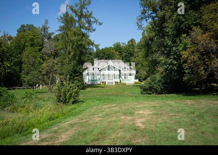 Una grande casa con un tetto verde si trova in un lussureggiante campo verde. La casa è circondata da alberi e presenta una finitura bianca. La scena è tranquilla e serena Foto Stock