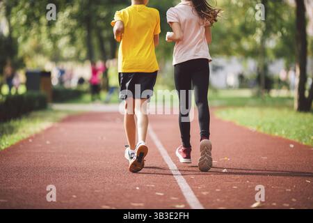 Primo piano delle gambe in allenatori di due bambini sulla pista da corsa dello stadio, vista posteriore. Ragazzo e ragazza sulla pista da jogging rossa nella corsa al parco. Gli scolari sono dentro Foto Stock