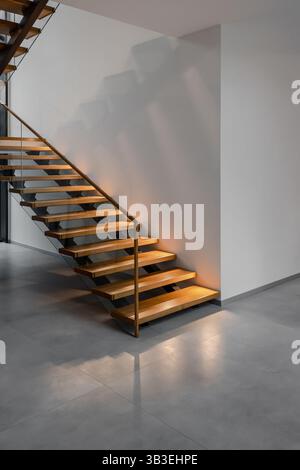 Scala vuota illuminata in legno in un edificio con pareti bianche e pavimento in cemento. Scala in legno con corrimano in una casa. Foto Stock