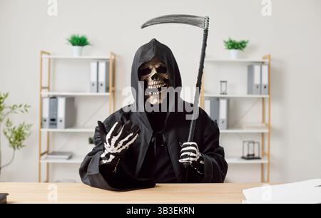 Triste Mietitore in un abito con cappuccio nero che tiene in mano una falce e si siede a una scrivania dell'ufficio Foto Stock