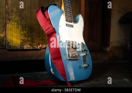 Chitarra elettrica blu con cinturino rosso appoggiato sul pavimento, inondata di luce naturale. Ideale per musica, stile di vita o progetti a tema vintage. Foto Stock