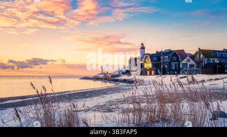 Mentre il sole tramonta su una tranquilla spiaggia dei Paesi Bassi, le case colorate riflettono sfumature dorate. Onde morbide baciano la spiaggia sabbiosa, creando un'atmosfera tranquilla al faro di Urk in inverno Foto Stock
