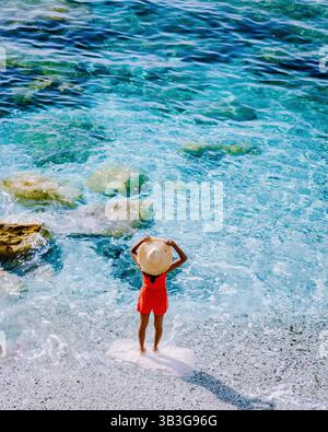 Sdraio e ombrelloni fiancheggiano la spiaggia di sabbia di Fegina a Monterosso, Italia. Una donna con un cappello da sole gode delle onde tranquille mentre il mare cristallino invita al relax. Foto Stock
